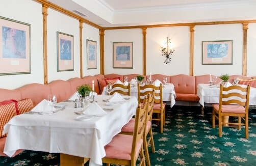 Ein Restaurant mit weiß gedeckten Tischen, Holzstühlen, rosa gepolsterten Bänken, gerahmter Kunst an den Wänden und einem grünen Blumenteppich.