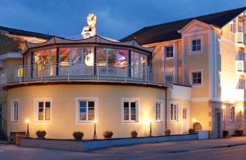 Ein zweistöckiges gelbes Gebäude mit großen Fenstern, einer überdachten Terrasse im Obergeschoss und sanfter Außenbeleuchtung in der Abenddämmerung.