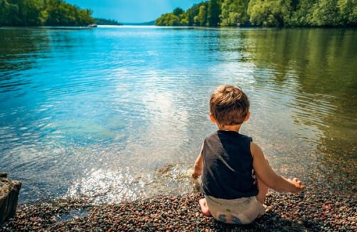 Ein kleines Kind sitzt an einem felsigen Ufer mit Blick auf einen ruhigen See, umgeben von Bäumen unter einem strahlend blauen Himmel.