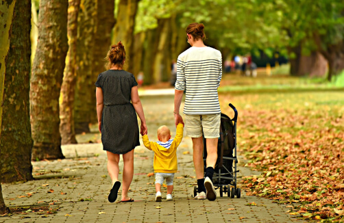Zwei Erwachsene und ein Kleinkind gehen einen von Bäumen gesäumten Weg entlang und halten das Kind an der Hand. Beide Erwachsenen sind leger gekleidet, und ein Kinderwagen steht in der Nähe. Blätter liegen auf dem Boden verstreut.