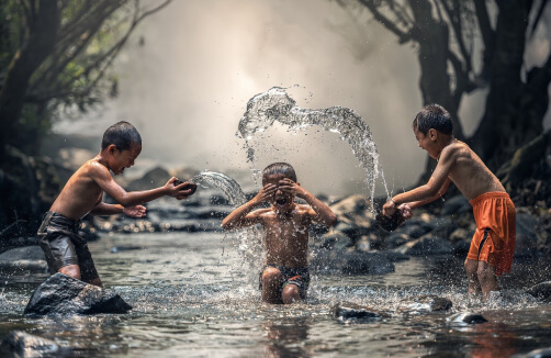 Drei Jungen spielen in einem seichten Bach und bespritzen sich gegenseitig mit Wasser zwischen Steinen und Bäumen.