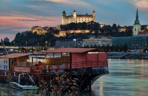 Ein Flussschiff mit der Aufschrift "Bratislava" legt bei Sonnenuntergang vor der beleuchteten Burg von Bratislava auf einem Hügel an, in der Nähe befinden sich Gebäude der Stadt und eine Kirchturmspitze.