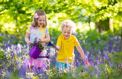 Zwei kleine Kinder stehen in einem Feld mit lila Blumen; ein Mädchen hält eine Gießkanne und Blumen, während ein Junge neben ihr einen kleinen Spaten hält.