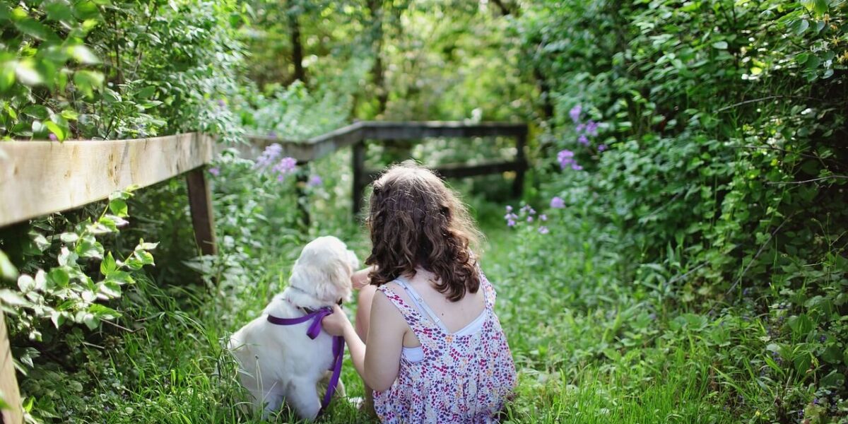 Ein Mädchen in einem geblümten Kleid sitzt im Gras neben einem weißen Hund mit einer lila Leine, umgeben von grünem Laub und Blumen.