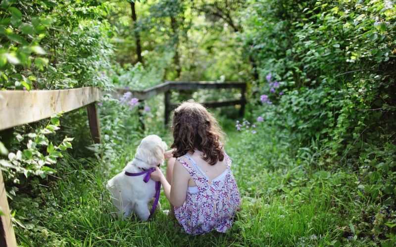 Ein Mädchen in einem geblümten Kleid sitzt im Gras neben einem weißen Hund mit einer lila Leine, umgeben von grünem Laub und Blumen.