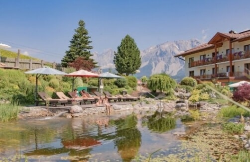Liegestühle und Sonnenschirme stehen neben einem natürlichen Teich in einem angelegten Garten, mit einem Holzgebäude und einer Bergkette im Hintergrund unter einem klaren Himmel.