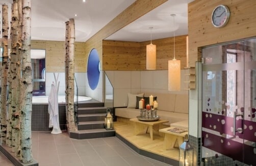 Moderne Spa-Lounge mit Birkendekor, Holzwänden, Kerzen, einer Uhr und einem Whirlpool-Bereich, der durch eine Treppe und eine Glasscheibe abgetrennt ist.