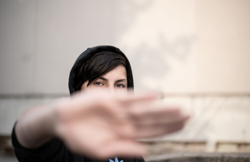 Eine Person in einem schwarzen Kapuzenpulli hält ihre Hand in Richtung Kamera, wobei sie ihr Gesicht teilweise verdeckt, vor einem neutralen Hintergrund im Freien.