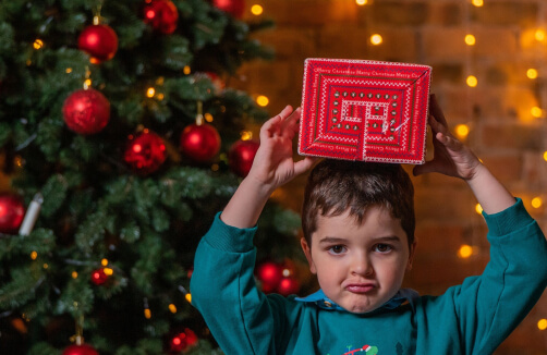 Ein kleiner Junge hält einen roten Geschenkkarton über seinem Kopf vor einem geschmückten Weihnachtsbaum mit roten Ornamenten und Lichterketten.