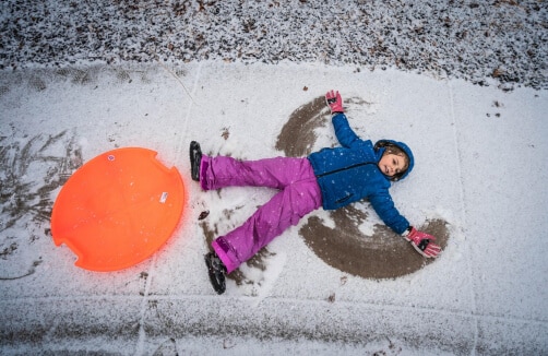 Ein Kind in blauer Jacke und rosa Hose macht einen Schneeengel auf einer dünnen Schneeschicht neben einem orangefarbenen Schlitten.