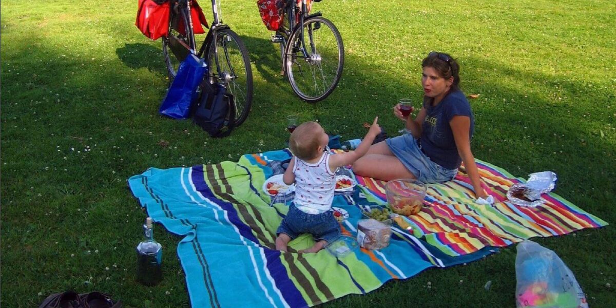 Eine Frau und ein kleines Kind sitzen auf einer gestreiften Decke und machen ein Picknick im Gras, in der Nähe stehen zwei Fahrräder, Essensbehälter und Spielzeug.