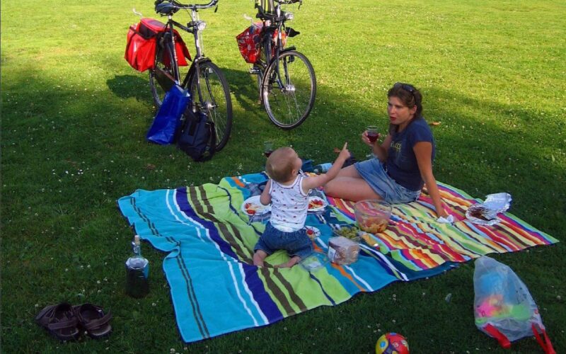 Eine Frau und ein kleines Kind sitzen auf einer gestreiften Decke und machen ein Picknick im Gras, in der Nähe stehen zwei Fahrräder, Essensbehälter und Spielzeug.