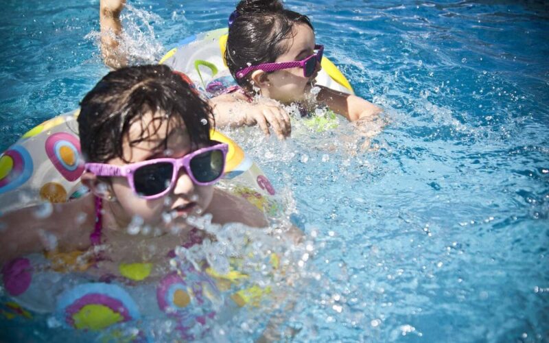 Zwei Kinder mit Sonnenbrillen schwimmen in einem Pool auf aufblasbaren Ringen und bespritzen das Wasser, während sie spielen.
