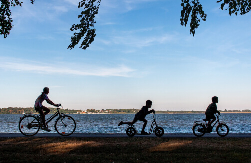 Drei Kinder fahren mit einem Fahrrad, einem Roller und einem kleinen Fahrrad unter freiem Himmel einen Uferweg entlang.