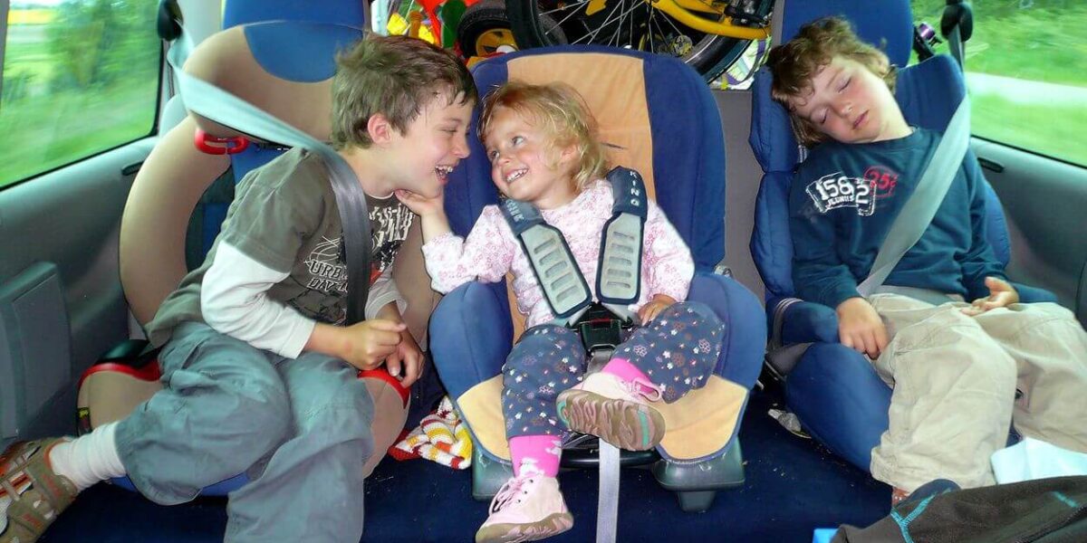 Drei Kinder sitzen auf dem Rücksitz eines Autos: zwei Jungen, einer schläft, der andere lächelt ein kleines Mädchen in einem Kindersitz an, hinter ihnen stehen Fahrräder und Spielzeug.