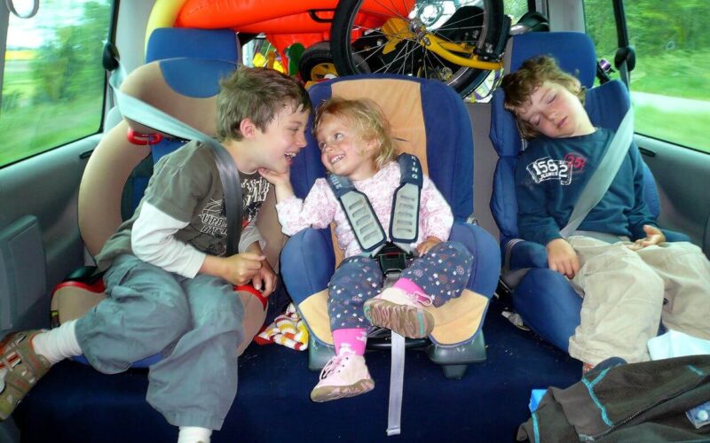 Drei Kinder sitzen auf dem Rücksitz eines Autos: zwei Jungen, einer schläft, der andere lächelt ein kleines Mädchen in einem Kindersitz an, hinter ihnen stehen Fahrräder und Spielzeug.