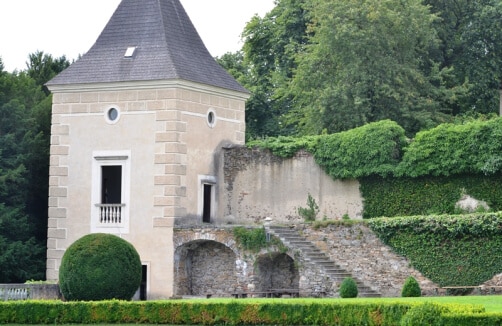 Ein steinernes Gebäude mit Ziegeldach steht neben einer mit Weinreben bewachsenen Mauer mit einer Steintreppe im Freien und beschnittenen Sträuchern davor.