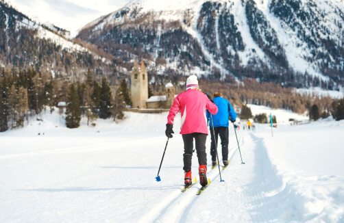 Zwei Personen beim Skilanglauf auf einer präparierten, verschneiten Loipe mit Bergen und einer Kirche im Hintergrund.