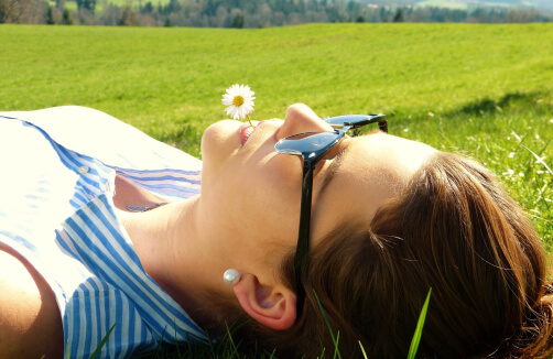 Eine im Gras liegende Frau mit Sonnenbrille, die ein Gänseblümchen im Mund hält, mit grünen Feldern und Bäumen im Hintergrund.
