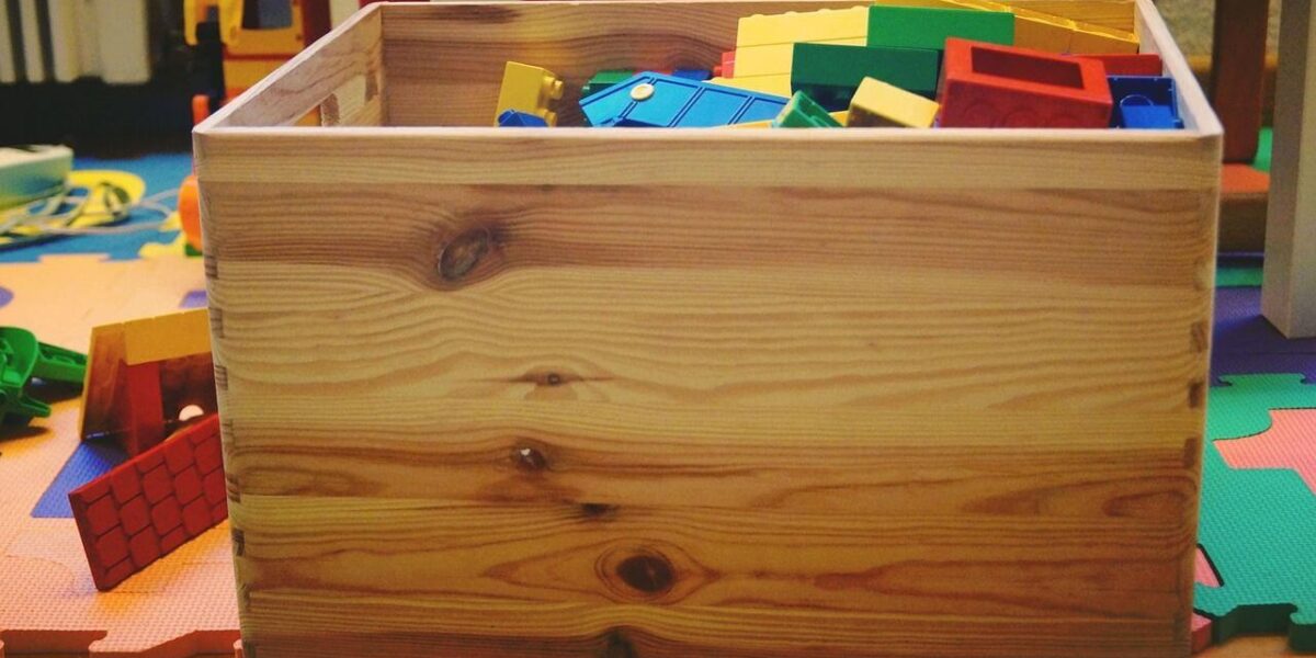 Eine Holzkiste, gefüllt mit bunten Plastikbausteinen, steht auf einer Spielmatte, umgeben von verstreutem Spielzeug.