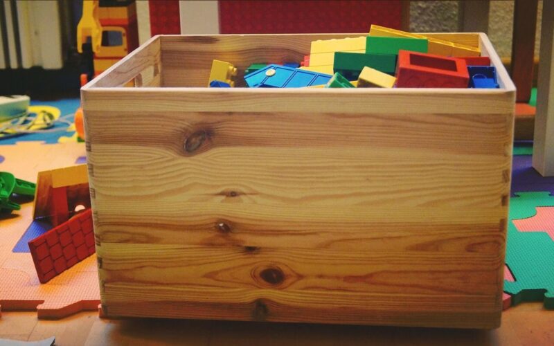 Eine Holzkiste, gefüllt mit bunten Plastikbausteinen, steht auf einer Spielmatte, umgeben von verstreutem Spielzeug.