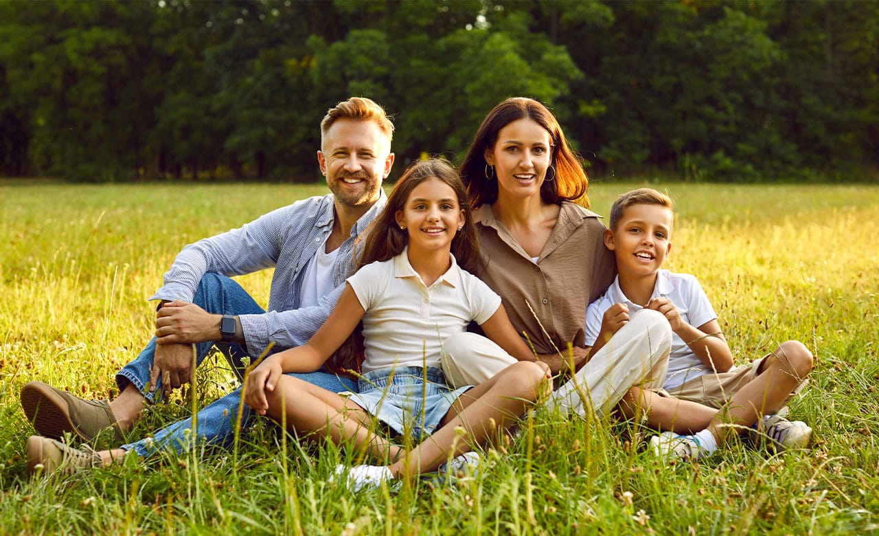 Eine vierköpfige Familie sitzt im Gras auf einem Feld und lächelt in die Kamera, während im Hintergrund Bäume zu sehen sind.