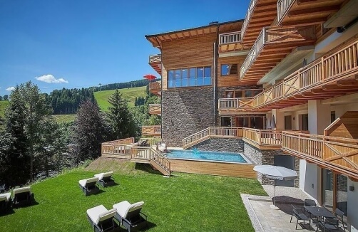 Modernes Hotelgebäude aus Holz und Stein mit Balkonen, Außenpool und Liegestühlen auf einer Rasenfläche, umgeben von Bäumen und Hügeln unter einem klaren blauen Himmel.