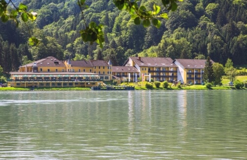Ein großer Hotelkomplex mit gelben Gebäuden steht an einem ruhigen See, umgeben von grünen, bewaldeten Hügeln im Tageslicht.