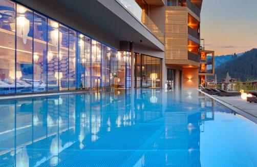 Ein modernes Hotel mit einem Infinity-Pool im Freien bei Sonnenuntergang, mit Glaswänden, Holzbalkonen und Liegestühlen am Rande des Pools.