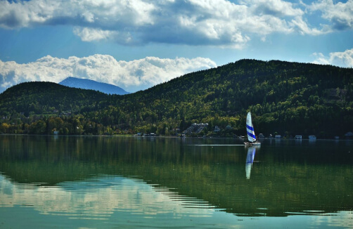 Ein Segelboot mit blauen und weißen Segeln fährt über einen ruhigen See, umgeben von grünen Hügeln und Bäumen, mit Wolken und Bergen im Hintergrund.