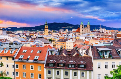 Blick auf eine europäische Stadtlandschaft mit farbenfrohen Gebäuden, Kirchtürmen und entfernten Hügeln unter einem leuchtenden, bewölkten Himmel bei Sonnenuntergang.