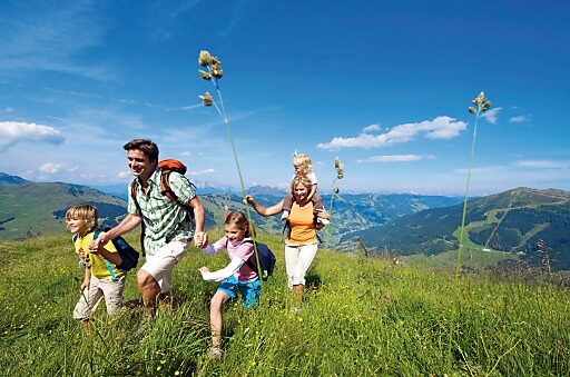 Eine fünfköpfige Familie, darunter drei Kinder, wandert an einem sonnigen Tag gemeinsam über eine Wiese, während im Hintergrund die Berge zu sehen sind.