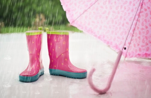 Zwei rosafarbene Regenstiefel stehen neben einem rosafarbenen Regenschirm auf einer nassen Fläche während eines Regenschauers, wobei das Grün im Hintergrund verschwimmt.