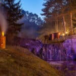Ein kleiner Wasserfall wird in der Abenddämmerung neben einem Waldstück mit violettem Licht beleuchtet; zwei Holzfackeln mit Flammen stehen auf dem Gras im Vordergrund.