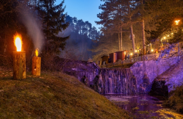 Ein kleiner Wasserfall wird in der Abenddämmerung neben einem Waldstück mit violettem Licht beleuchtet; zwei Holzfackeln mit Flammen stehen auf dem Gras im Vordergrund.