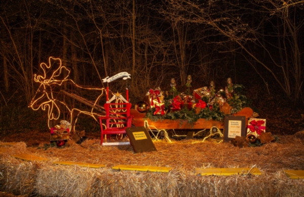 Eine Weihnachtsausstellung zeigt ein beleuchtetes Rentier und einen Schlitten mit eingepackten Geschenken, Grünzeug und Schleifen, die auf Heuballen mit Bäumen im Hintergrund stehen.