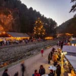 In der Abenddämmerung spazieren die Menschen zwischen den Ständen eines Weihnachtsmarktes in der Nähe eines bewaldeten Hügels, wo festliche Lichter und ein großer geschmückter Baum zu sehen sind.