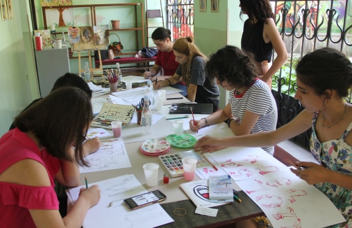 Eine Gruppe von Menschen sitzt in einem Kunstatelier um einen Tisch herum und zeichnet und malt auf Papier, wobei verschiedene Kunstmaterialien auf dem Tisch verstreut sind.