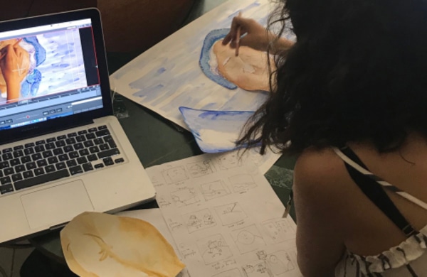 Eine Person sitzt an einem Tisch und skizziert und malt, während sie ein Animationsvideo auf einem Laptop betrachtet, auf dem Storyboards und Kunstwerke ausgebreitet sind.