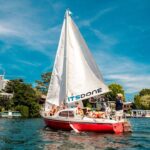 Eine Gruppe von Menschen segelt mit einem rot-weißen Segelboot mit der Aufschrift "ITSDONE" auf einem ruhigen Gewässer mit Gebäuden und Bäumen im Hintergrund unter blauem Himmel.