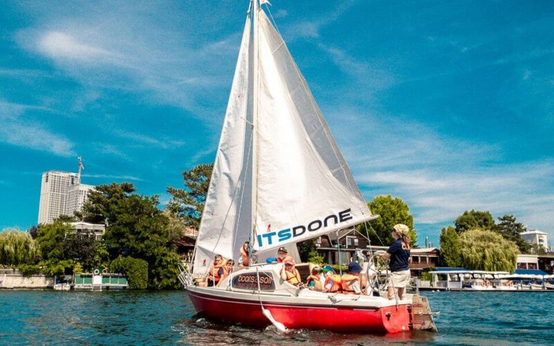 Eine Gruppe von Menschen segelt mit einem rot-weißen Segelboot mit der Aufschrift "ITSDONE" auf einem ruhigen Gewässer mit Gebäuden und Bäumen im Hintergrund unter blauem Himmel.