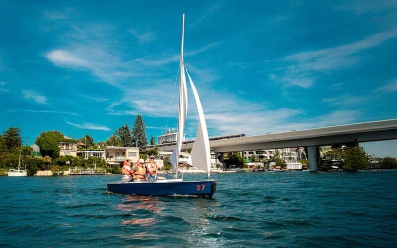 Eine kleine Gruppe von Personen, die Schwimmwesten tragen, segelt auf einem blauen Segelboot auf einem See in der Nähe einer Brücke und von Häusern am Ufer unter einem klaren blauen Himmel.