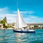 Zwei Personen segeln an einem sonnigen Tag auf einem kleinen blau-weißen Segelboot, im Hintergrund sind Häuser und eine Brücke zu sehen.