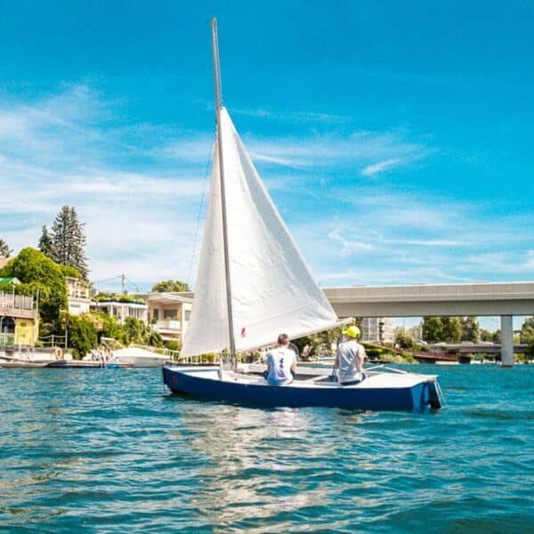 Zwei Personen segeln an einem sonnigen Tag auf einem kleinen blau-weißen Segelboot, im Hintergrund sind Häuser und eine Brücke zu sehen.