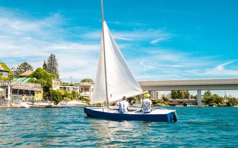 Zwei Personen segeln an einem sonnigen Tag auf einem kleinen blau-weißen Segelboot, im Hintergrund sind Häuser und eine Brücke zu sehen.