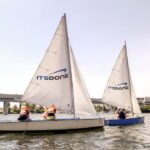 Auf dem Wasser sind zwei Segelboote mit der Aufschrift "IT'S DONE" zu sehen, auf denen jeweils Personen mit orangefarbenen Schwimmwesten sitzen. Im Hintergrund sind eine Brücke und Gebäude zu sehen.