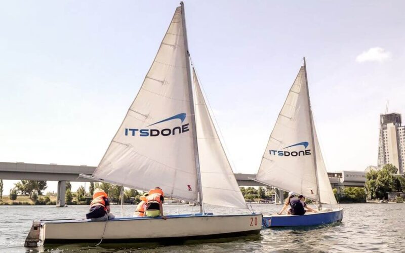Auf dem Wasser sind zwei Segelboote mit der Aufschrift "IT'S DONE" zu sehen, auf denen jeweils Personen mit orangefarbenen Schwimmwesten sitzen. Im Hintergrund sind eine Brücke und Gebäude zu sehen.