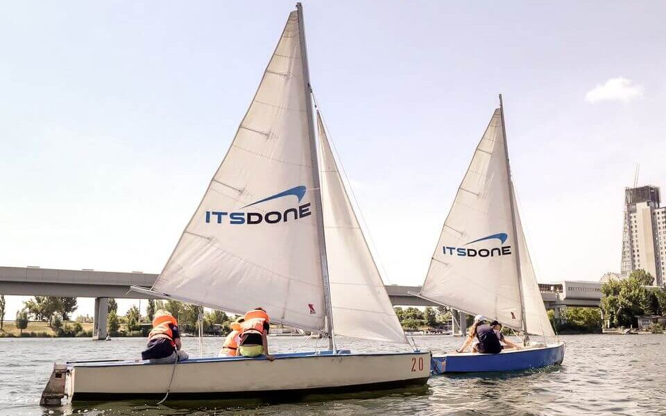 Auf dem Wasser sind zwei Segelboote mit der Aufschrift "IT'S DONE" zu sehen, auf denen jeweils Personen mit orangefarbenen Schwimmwesten sitzen. Im Hintergrund sind eine Brücke und Gebäude zu sehen.