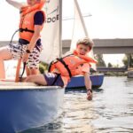 Zwei Kinder mit orangefarbenen Schwimmwesten sitzen auf einem kleinen Segelboot; eines lehnt sich über den Rand und berührt mit seiner Hand das Wasser.