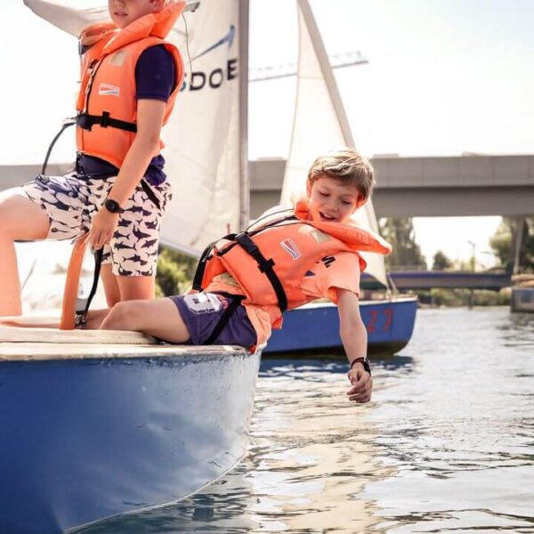 Zwei Kinder mit orangefarbenen Schwimmwesten sitzen auf einem kleinen Segelboot; eines lehnt sich über den Rand und berührt mit seiner Hand das Wasser.